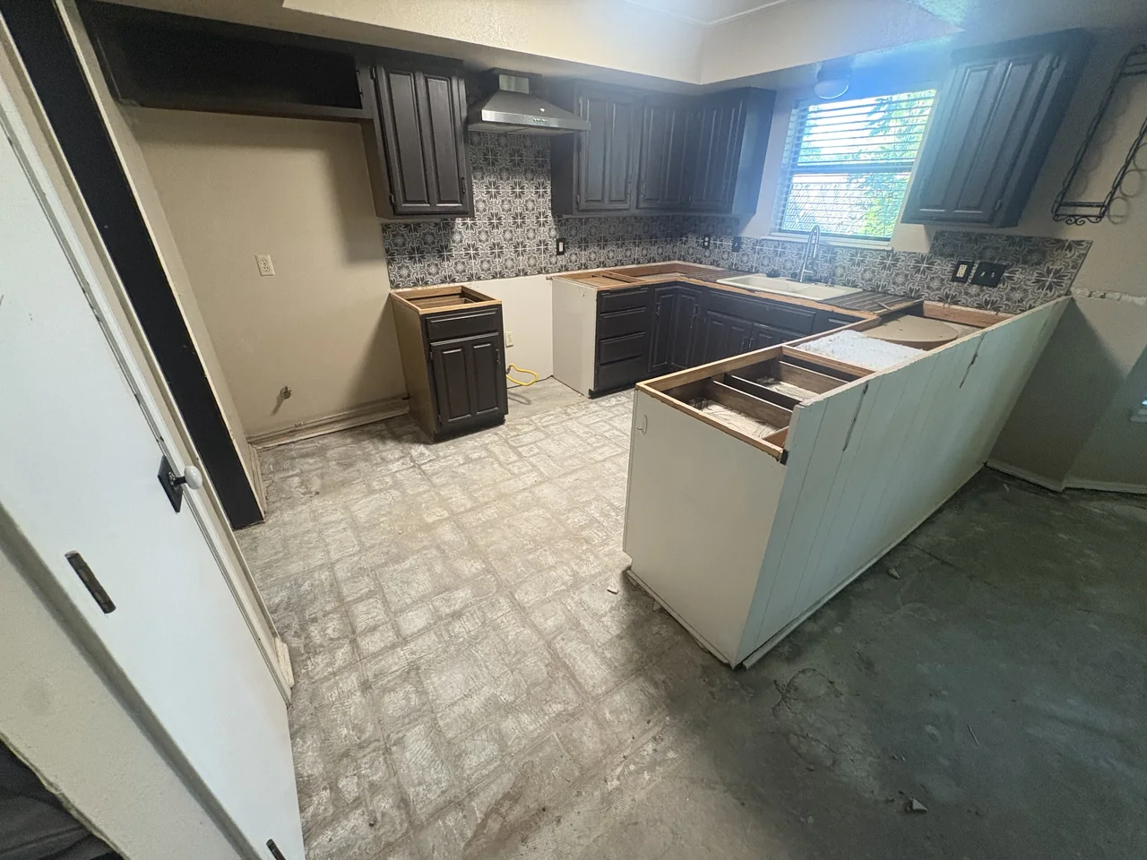 Kitchen before renovation, with dark dated cabinets, torn-out countertops and old vinyl tile.