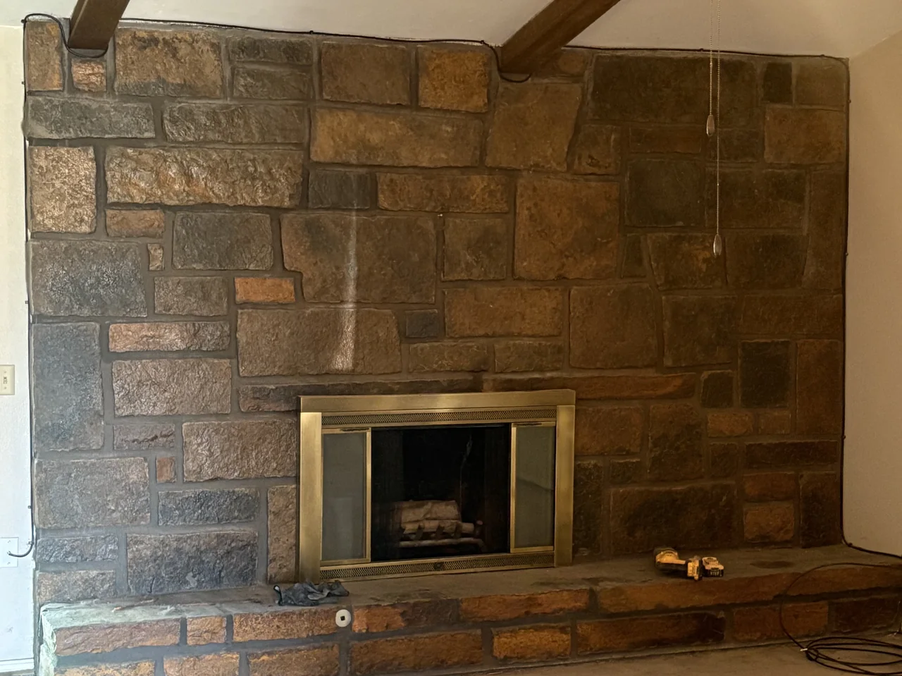 Living room before renovation, with a dark dated stone fireplace, brass doors and bare concrete floor.
